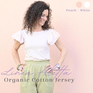 Puede incluir: Una camiseta blanca de punto de algodón orgánico con mangas con volantes y pantalones verde claro. El texto "Linea Aletta" y "Organic Cotton Jersey" son visibles. El fondo es de color melocotón claro.