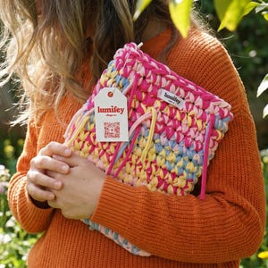 May include: A colorful, handmade clutch with a tag that reads "lumifey". The clutch is made of woven fabric in shades of pink, yellow, blue, and white. It has a rectangular shape and is held in front of a person wearing an orange sweater.