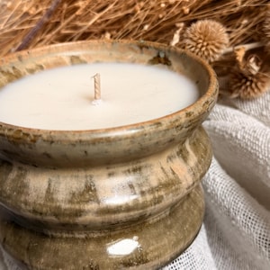 Peut inclure: Une bougie artisanale en céramique de conception rustique empilée. La bougie a une cire blanche crémeuse et une seule mèche. Le récipient en céramique est verni dans des tons bruns et verts, avec une finition texturée. Des plantes séchées sont en arrière-plan.