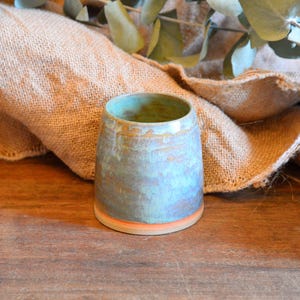 Peut inclure: Un gobelet en céramique fait à la main avec un glaçage bleu et vert, rehaussé de teintes orange et marron. La petite tasse cylindrique repose sur une surface en bois, avec un tissu de jute et de la verdure en arrière-plan. La poterie a une esthétique rustique et artisanale.