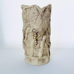 Puede incluir: Un jarrón cilíndrico beige con una apariencia texturizada similar a la piedra. El jarrón presenta un relieve detallado de una gran hoja de uva y un racimo de uvas. El diseño está tallado en la superficie, creando una estética rústica y natural.