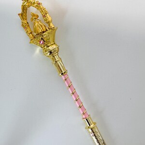Puede incluir: Un cetro decorativo dorado con un mango rosa. El cetro presenta una pequeña figura con un vestido en la parte superior, rodeada por un marco dorado con gemas rosas. El mango es rosa con detalles dorados. El cetro mide unos 30 cm de largo.