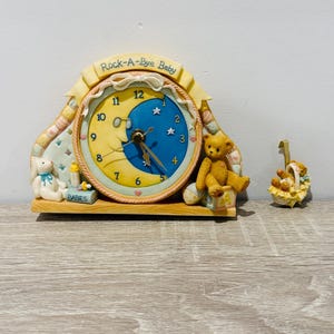 May include: A decorative clock featuring a yellow crescent moon face with blue night sky, labelled Rock-A-Bye Baby. The clock includes a white rabbit, a baby bottle, a book labelled BABIES, and a brown teddy bear. A small matching bear ornament sits nearby.
