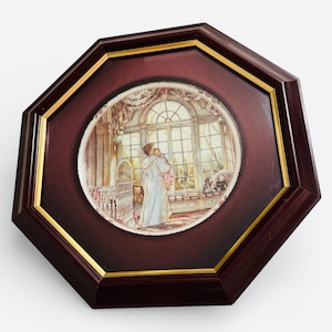 May include: An octagonal framed decorative plate. The plate features a detailed illustration of a person holding a baby near a large window. The frame is dark brown with a gold inner trim. The plate's illustration has soft, muted colours.