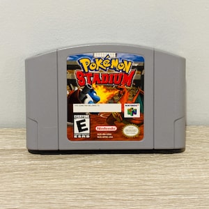 Puede incluir: Un cartucho de juego gris de Nintendo 64 para Pokémon Stadium. La etiqueta muestra a Blastoise y Charizard en un estadio, el logotipo de Pokémon Stadium, una clasificación ESRB E, el logo de Nintendo 64 y el texto This Game Pak Belongs To.