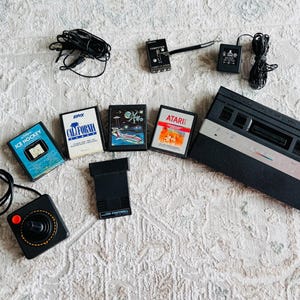 Könnte beinhalten: Eine Sammlung von Atari-Videospielkassetten und Zubehör aus der Vintage-Ära, darunter eine schwarze Konsole, ein Joystick und Spielkassetten mit Titeln wie "Ice Hockey" und "California". Die Artikel sind auf einer strukturierten weißen Oberfläche angeordnet.