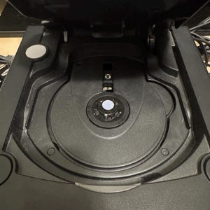 Könnte beinhalten: Eine schwarze Videospielkonsole mit geöffnetem Laufwerksdeckel. Der Top-Loading-Mechanismus verfügt über eine zentrale kreisförmige Spindel zur Aufnahme von Discs, eine runde Auswurftaste auf der linken Seite und ein mattes schwarzes Kunststoffgehäuse.