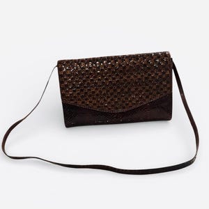 May include: A brown, rectangular shoulder bag with a textured, mosaic-like pattern on the flap. The bag has a smooth, dark brown base and a thin, matching strap. The bag is set against a white background.