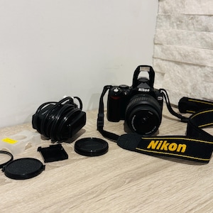 May include: A black Nikon DSLR camera with an attached lens and a black and yellow branded neck strap. Included are a black power cable, a translucent plastic lens cap holder, two circular black lens caps, and a small black square accessory cover.