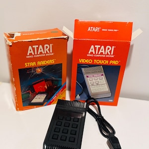 May include: A black Atari Video Touch Pad controller with a wired connector, accompanied by its original orange cardboard packaging for the Star Raiders game and the Video Touch Pad accessory, both featuring the Atari Video Computer System logo.
