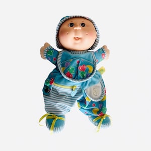 Könnte beinhalten: Eine Vintage-Cabbage-Patch-Kid-Puppe in einem blauen Outfit mit passender Kapuze. Das Outfit der Puppe hat bunte Stern- und Herzmuster, gestreifte Hosen und gelbe Schleifenakzente. Die Puppe hat rosige Wangen und einen lächelnden Ausdruck.