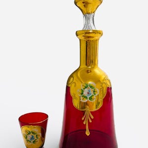 Puede incluir: Juego de decantador y vaso de chupito de cristal rojo rubí con detalles dorados. El decantador tiene un tapón dorado y está decorado con diseños florales pintados a mano. El vaso de chupito refleja el diseño del decantador.