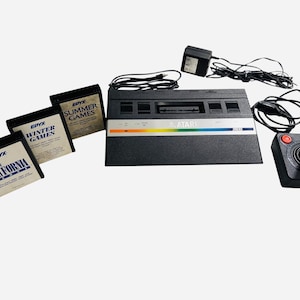 May include: A classic Atari 2600 video game console, joystick, and power supply. The black console has a colourful stripe. Three game cartridges are visible, labelled "California", "Winter Games", and "Summer Games".