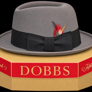 May include: A grey fedora hat with a black grosgrain ribbon band and a small red, black, and white feather accent. The hat rests on a tan octagonal box with red trim, featuring the text DOBBS, New York, and Fifth Avenue printed on the sides.