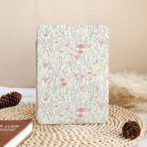 May include: A floral patterned tablet case featuring a white background with a delicate design of pink, yellow, and orange flowers and green stems. The case is rectangular and appears to be made of a smooth material. The design is reminiscent of a spring meadow.