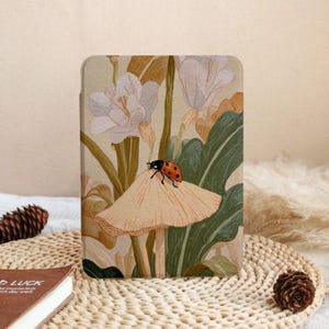 Puede incluir: Una funda rectangular para tableta con una ilustración botánica de una mariquita roja con puntos negros posada sobre un hongo de color tostado claro. El diseño incluye flores blancas, hojas verdes y tallos tostados sobre un fondo crema.