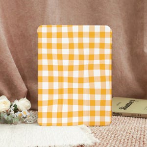 Könnte beinhalten: Eine Tablet-Hülle mit einem gelb-weißen Vichy-Muster. Die Hülle hat eine rechteckige Form mit abgerundeten Ecken. Im Hintergrund ist ein Buch mit dem Titel "Butter" zu sehen.