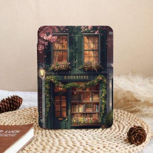 Può includere: Una custodia rettangolare per e-reader con un'illustrazione stampata di una facciata di una libreria verde scuro con grandi vetrate, scaffali pieni di libri, ghirlande floreali pendenti e fiori di ciliegio rosa su un edificio in mattoni.