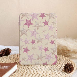 Könnte beinhalten: Eine beige Tablet-Hülle mit einem Muster aus rosa und silbernen Glitzersternen. Die Sterne sind unterschiedlich groß und über die Oberfläche verteilt. Die Hülle ist rechteckig und scheint aus einem strapazierfähigen Material zu bestehen.
