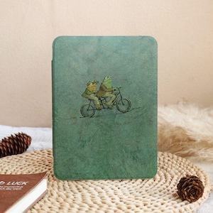 May include: A green tablet case featuring an illustration of two frogs riding a bicycle. The frogs are wearing hats and jackets, set against a textured, mottled green background. The case has rounded corners.