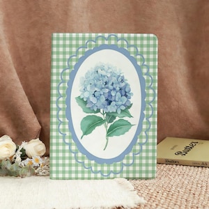 May include: A notebook with a green and white gingham pattern. The cover features a blue oval frame with a watercolor illustration of a blue hydrangea flower. The notebook is rectangular and has a soft, floral aesthetic.