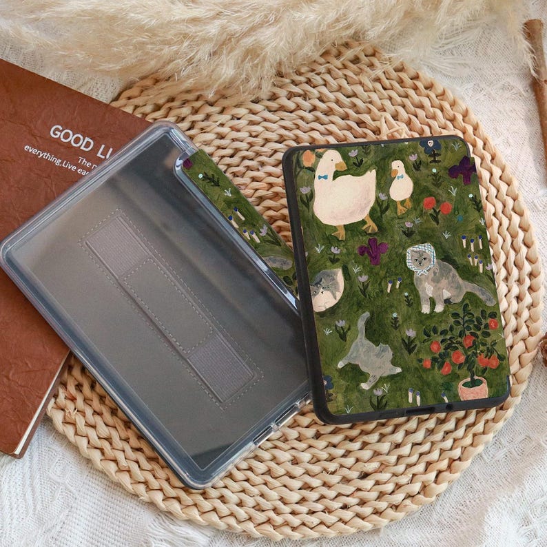 May include: A clear tablet case with a green cover featuring a whimsical design of geese, cats, and flowers on a green background. The case is open, revealing the interior. A brown book with the words "GOOD LI" is visible.