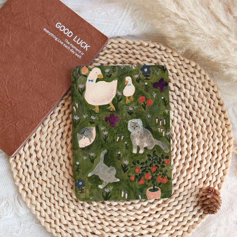 May include: A brown notebook with the text "GOOD LUCK" and a decorative cover featuring ducks, cats, and flowers on a green background. The notebook is placed on a woven mat.