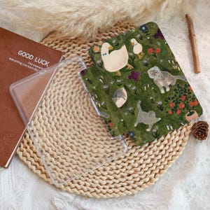 May include: A clear phone case and a patterned phone case featuring a whimsical design of geese and cats in a garden setting. The patterned case has a green background with floral and animal illustrations. A brown book with the text "GOOD LUCK" is also visible.