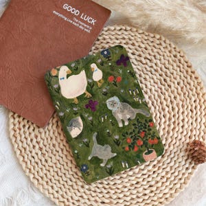 May include: A brown notebook with the words "GOOD LUCK" and "The moment is everything. Live each day well." A green notebook with illustrations of ducks, cats, and flowers. The notebooks are on a woven mat.