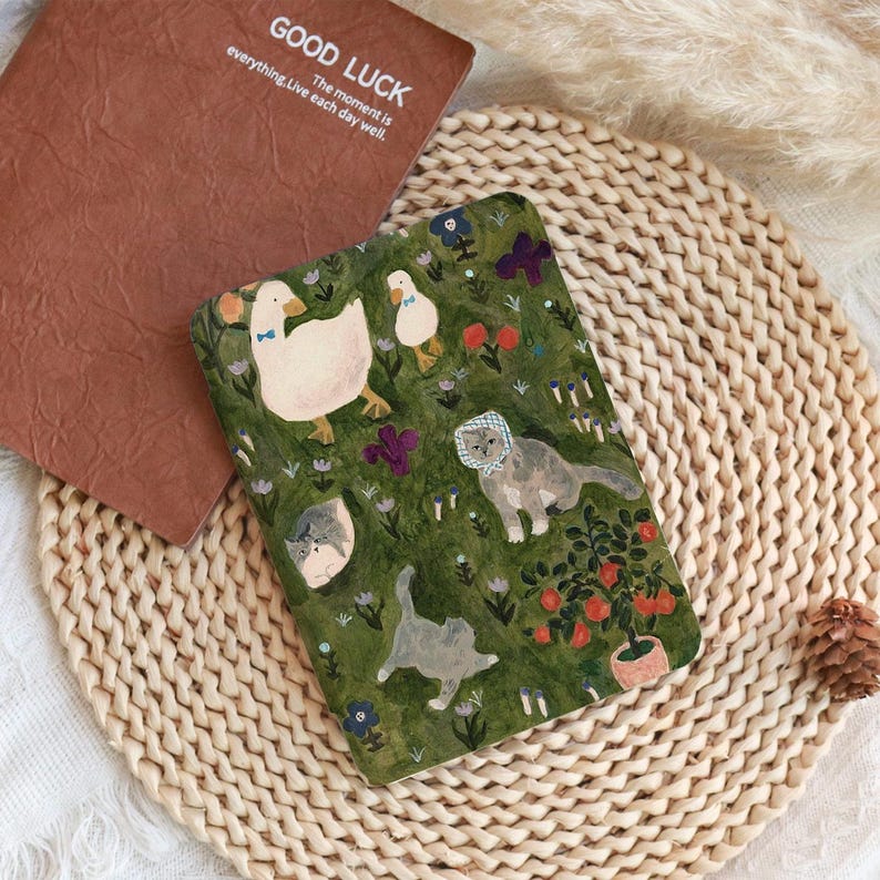May include: A brown notebook with the words "GOOD LUCK" and "The moment is everything. Live each day well." A green notebook with illustrations of ducks, cats, and flowers. The notebooks are on a woven mat.