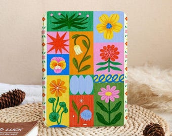 May include: A rectangular notebook with a colorful floral design. The cover features a grid of nine squares, each with a different flower illustration in vibrant colors like yellow, orange, pink, and blue. The notebook is resting on a woven mat.