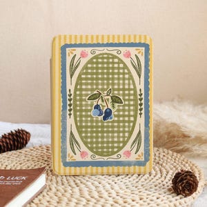 Puede incluir: Un diario decorativo con un borde a rayas amarillas y azules. El centro presenta un patrón de cuadros verdes con una ilustración de arándanos. Acentos florales rosas y un borde festoneado azul completan el diseño.