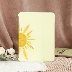 May include: A light yellow tablet case with a watercolor sun design and the text "the sun on the side out". The case is closed and has a smooth surface. A book labeled "Butter" is visible in the background.