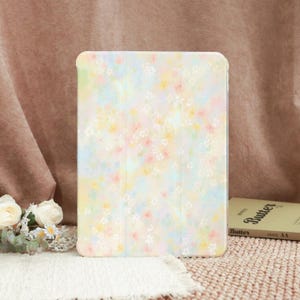 Op de afbeelding: Een pastelkleurige tablethoes met een bloemenpatroon. De hoes heeft een zacht, aquarelachtig ontwerp met kleine roze, gele en blauwe bloemen op een lichte achtergrond. De hoes is gesloten en lijkt gemaakt van een glad materiaal.