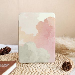 May include: A rectangular e-reader case featuring a soft watercolor-style print with abstract cloud-like shapes in muted shades of dusty pink, cream, and sage green. The surface has a smooth finish and rounded corners, displaying a gentle color gradient.