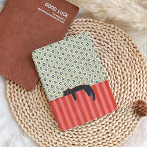 Puede incluir: Un cuaderno marr&oacute;n con el texto "GOOD LUCK" y un cuaderno estampado con un gato negro descansando sobre una secci&oacute;n a rayas rojas y rosas. La secci&oacute;n estampada tiene un dise&ntilde;o floral.
