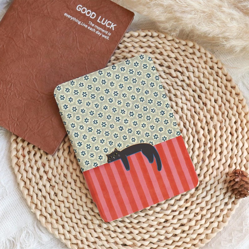 Puede incluir: Un cuaderno marr&oacute;n con el texto "GOOD LUCK" y un cuaderno estampado con un gato negro descansando sobre una secci&oacute;n a rayas rojas y rosas. La secci&oacute;n estampada tiene un dise&ntilde;o floral.