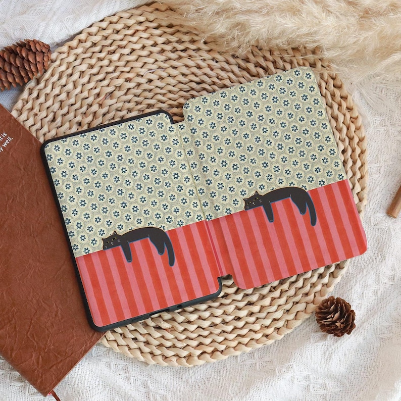 Puede incluir: Una funda negra para tableta con un dise&ntilde;o caprichoso. La funda presenta una secci&oacute;n a rayas rojas y rosas con dos ilustraciones de gatos negros. La parte superior tiene un patr&oacute;n floral repetitivo en verde y azul.