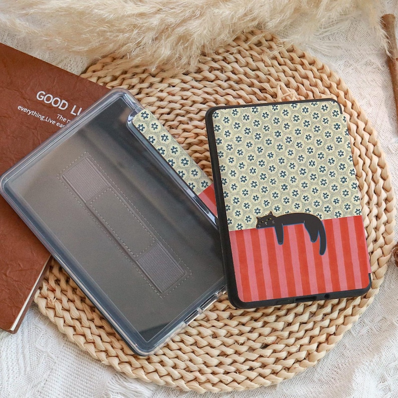 Puede incluir: Una funda para lector electr&oacute;nico y una funda protectora transparente. La funda presenta un gato negro sobre una secci&oacute;n a rayas rojas y rosas, con un estampado floral encima. Un libro marr&oacute;n con el texto "GOOD LI" es visible.