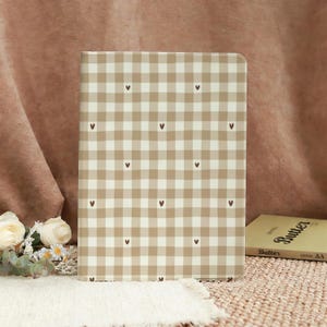 May include: A beige and white checkered notebook cover with small brown hearts scattered across the surface. The notebook is rectangular and stands upright. The background includes a book and flowers.