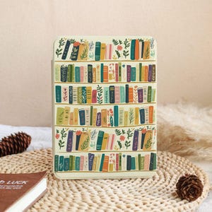 Peut inclure: Un étui crème avec un motif de bibliothèque colorée, avec des livres aux tons de vert, orange, bleu et violet, et des accents floraux. L'étui rectangulaire est probablement destiné à un appareil électronique.