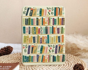 Puede incluir: Una funda de color crema con un diseño de estantería colorida, con libros en tonos verdes, naranjas, azules y morados, y detalles florales. La funda rectangular es probablemente para un dispositivo electrónico.