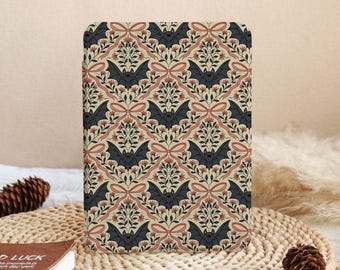 May include: A rectangular notebook with a repeating pattern of bats, bows, and floral elements in black, peach, and cream. The design has a vintage, gothic aesthetic. The notebook is displayed on a woven mat.
