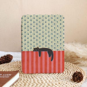 Puede incluir: Una funda rectangular con una ilustraci&oacute;n de un gato negro. La parte superior tiene un patr&oacute;n floral en beige y azul, mientras que la parte inferior tiene rayas verticales rojas. El gato est&aacute; acostado sobre las rayas rojas.