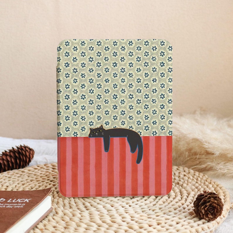 Puede incluir: Una funda rectangular con una ilustraci&oacute;n de un gato negro. La parte superior tiene un patr&oacute;n floral en beige y azul, mientras que la parte inferior tiene rayas verticales rojas. El gato est&aacute; acostado sobre las rayas rojas.