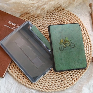 Może przedstawiać: Zielone etui na tablet z fantazyjną ilustracją dw&oacute;ch żab na rowerze. Etui jest otwarte, ukazując przezroczyste wnętrze i brązową książkę z napisem "GOOD LI".