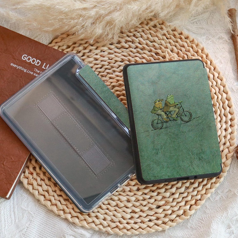 Może przedstawiać: Zielone etui na tablet z fantazyjną ilustracją dw&oacute;ch żab na rowerze. Etui jest otwarte, ukazując przezroczyste wnętrze i brązową książkę z napisem "GOOD LI".