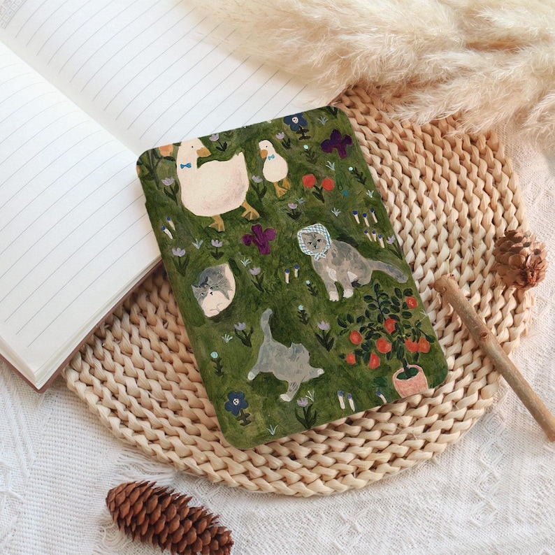 May include: A decorative journal with a whimsical illustration of geese and cats in a garden setting. The cover features a green background with colorful flowers and a potted plant. The journal is open, revealing lined pages.