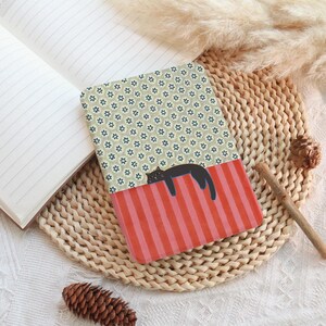 Puede incluir: Un cuaderno estampado con una ilustraci&oacute;n de un gato negro descansando sobre una secci&oacute;n a rayas rojas y rosas. El cuaderno tiene un dise&ntilde;o floral en la mitad superior y est&aacute; abierto para revelar p&aacute;ginas rayadas. El cuaderno est&aacute; sobre una estera tejida.