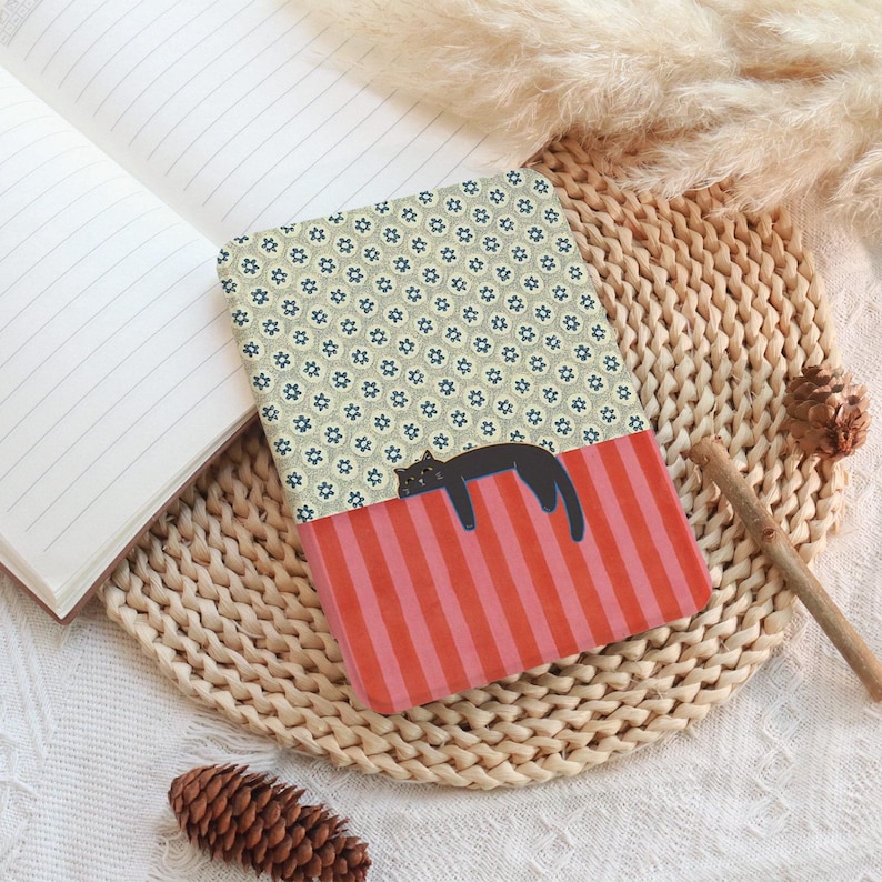 Puede incluir: Un cuaderno estampado con una ilustraci&oacute;n de un gato negro descansando sobre una secci&oacute;n a rayas rojas y rosas. El cuaderno tiene un dise&ntilde;o floral en la mitad superior y est&aacute; abierto para revelar p&aacute;ginas rayadas. El cuaderno est&aacute; sobre una estera tejida.